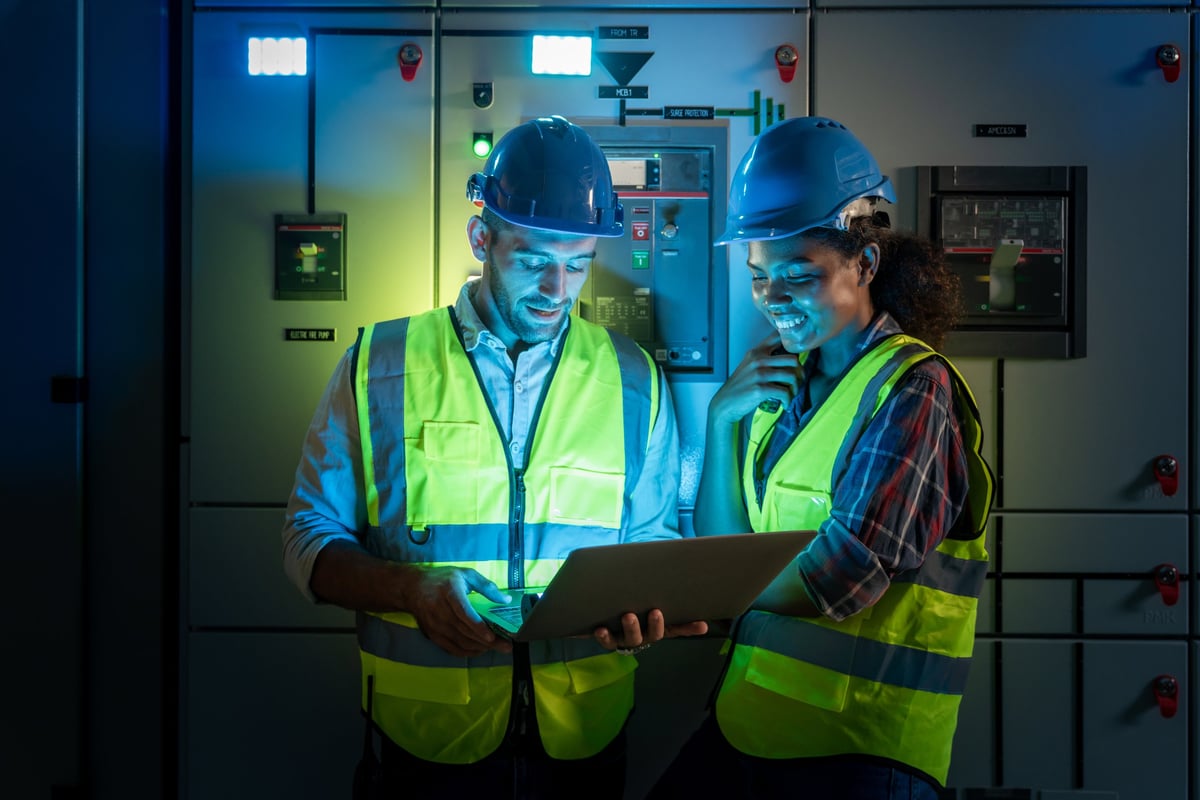 Electrical engineer use laptop to inspection and testing of electrical system in control room. Electrician and technician working night shifts at distribution board. Multi-Ethnic Blue-collar worker.