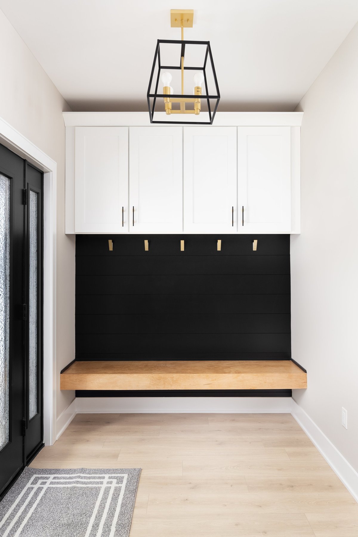A modern farmhouse entryway with bench seating, black shiplap, white cabinets, gold coat hooks, and black and gold light fixture.