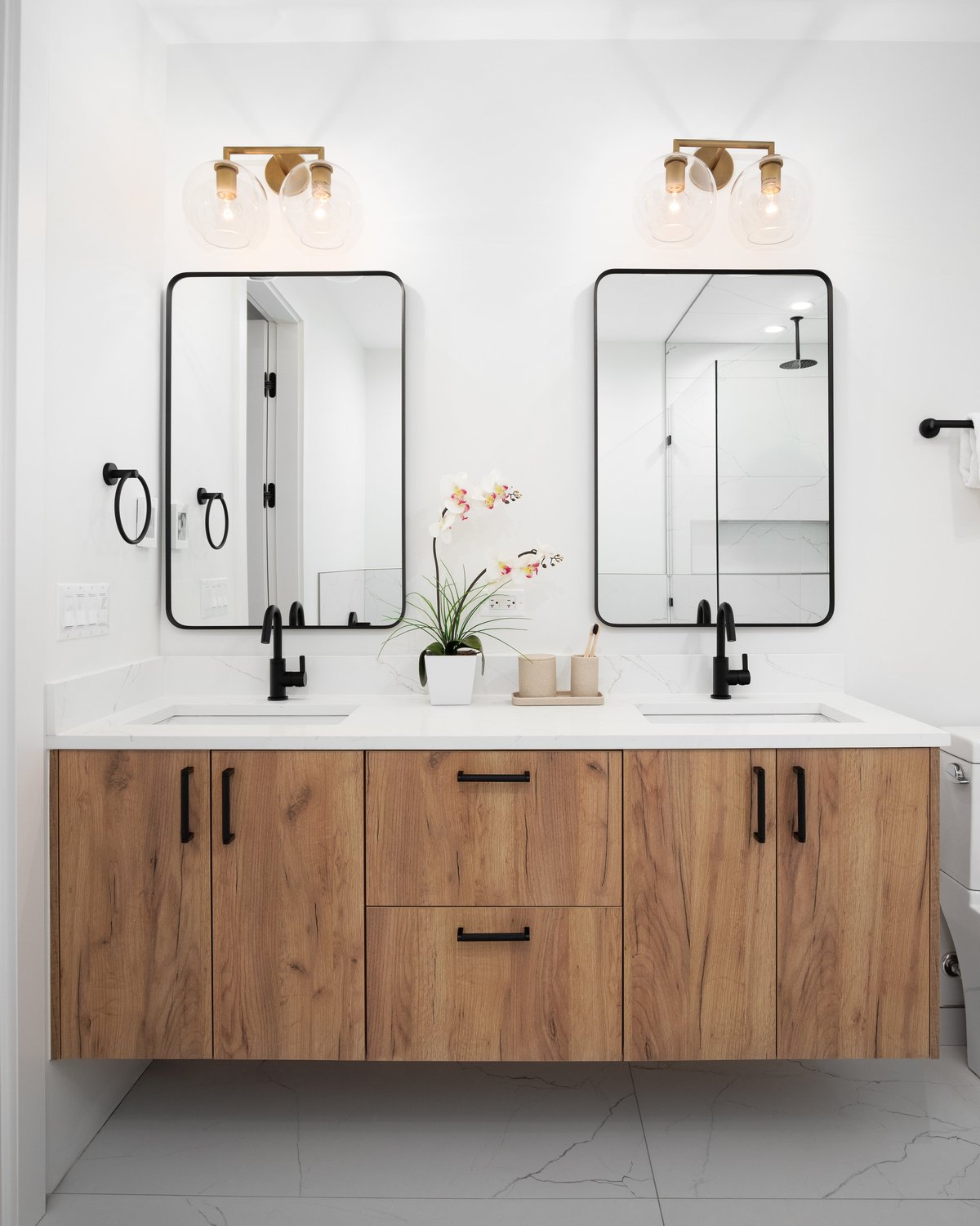 A bathroom with a floating wood vanity cabinet, decorations on the white marble countertop, gold lights mounted above two black mirrors, and black faucets