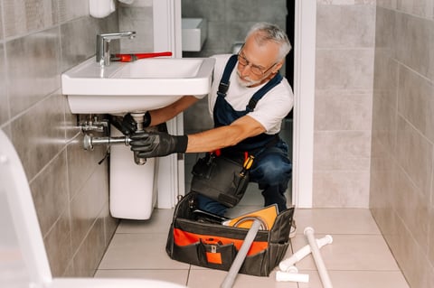 Plumber is working under a sink, installing the piping with a wrench, in a bathroom. His toolbox is open on the floor next to him