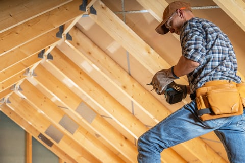 A construction worker is focused on installing wooden beams in an attic space during a home renovation. Visible are the wooden structures overhead, showcasing the progress of the project in bright, natural light.