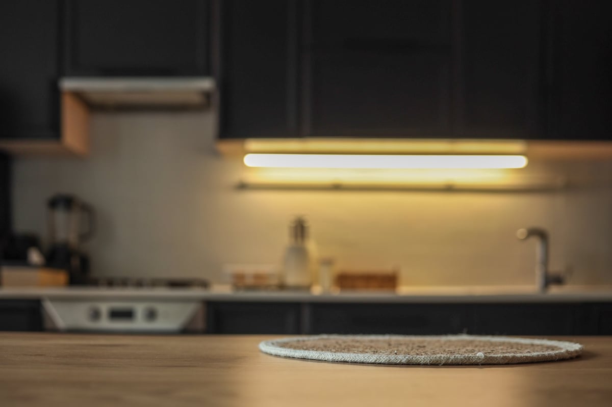 Cozy modern kitchen interior with a round textured placemat on a wooden table in soft focus foreground. Minimalist design with dark cabinets and under-cabinet lighting in the background.