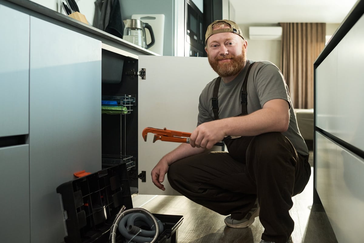 Caucasian middle aged man kneeling in modern kitchen holding pipe wrench and smiling at camera