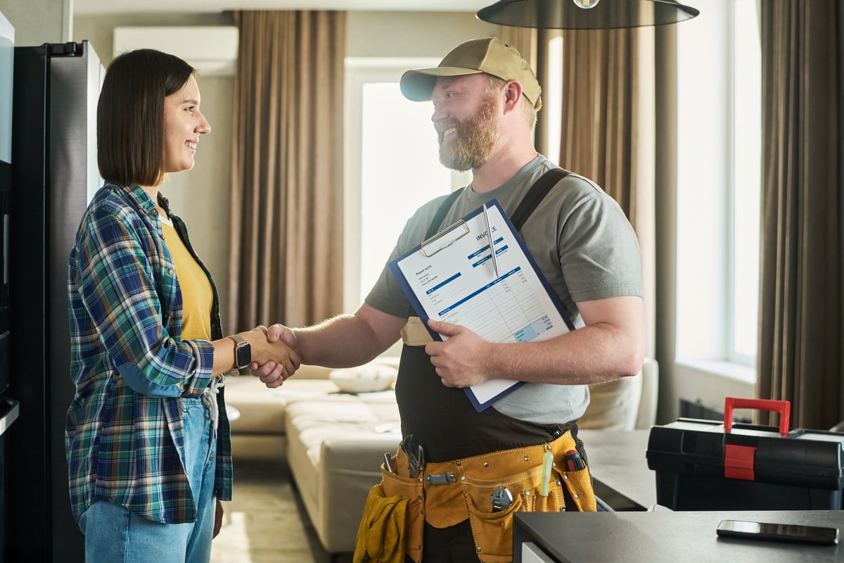 Caucasian young adult woman shaking hands with a middle aged male repairman holding a clipboard and invoice in a modern living room, both smiling