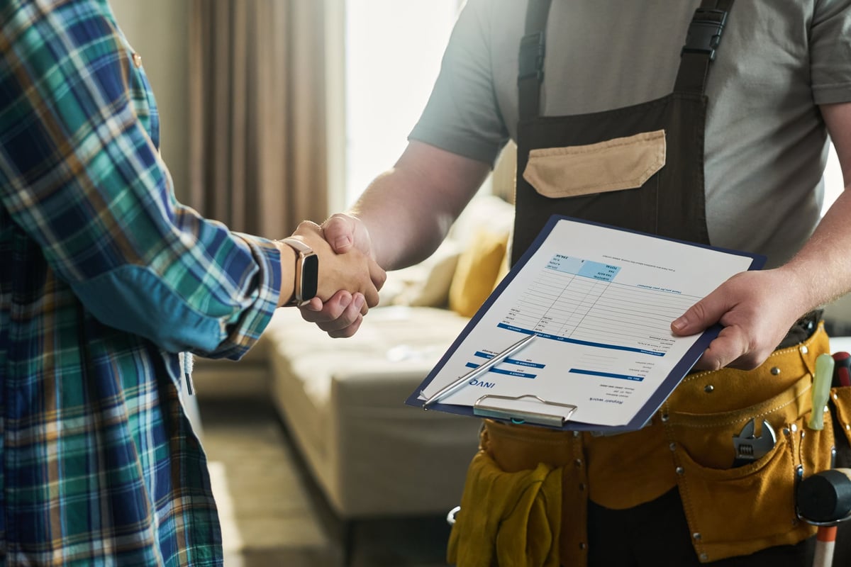 Caucasian middle aged man wearing tool belt holding invoice clipboard shaking hands with unseen young adult Caucasian woman in home interior, confirming service completion