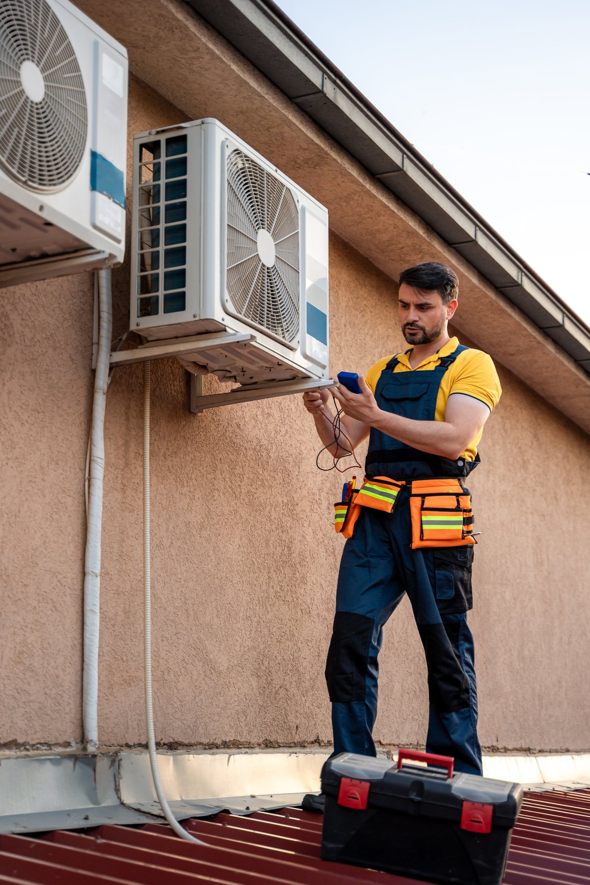 A technician stands on a rooftop using tools to inspect and maintain air conditioning units.