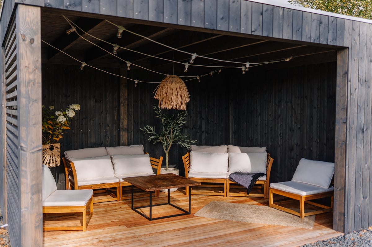 Modern outdoor pergola with cozy white-cushioned wooden seating, string lights, and potted plants for a stylish backyard retreat.