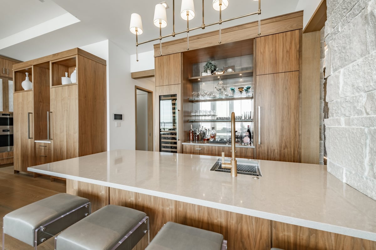 This image displays a modern, open concept kitchen, living room and bar area featuring a seamless flow between the kitchen bar and living room increasing natural light. Two large kitchen islands one with sink and seating for four, and extensive wood cabinetry that includes integrated appliances and glass fronted display cabinets. Countertops are white, light wood flooring and stone accents on pillars a fireplace wall and large floor to ceiling windows reveal a sunny summer day with blue sky. Several large spherical black and metal wrought iron chandeliers and natural light under beamed ceilings.