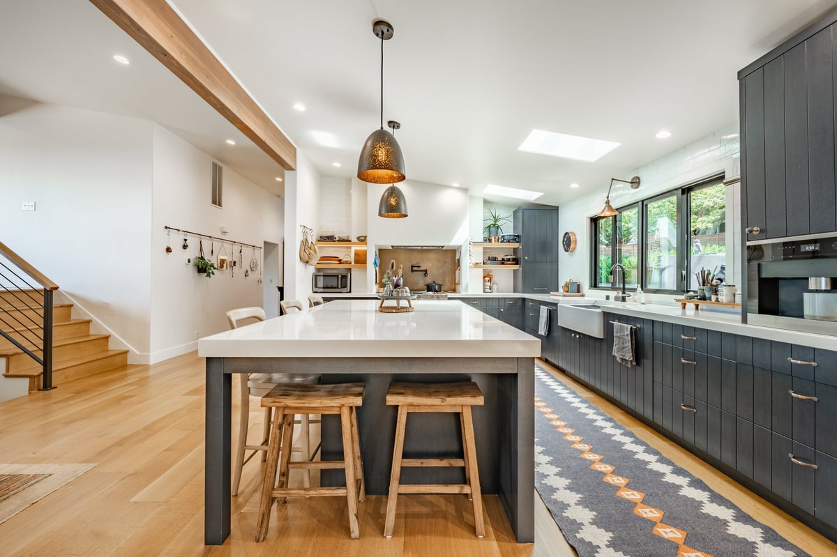 A modern kitchen with dark cabinetry light countertops warm wood flooring. Featuring a large central island with white countertop and seating for four using wooden stools. Two hammered metal pendant lights hang over the island.