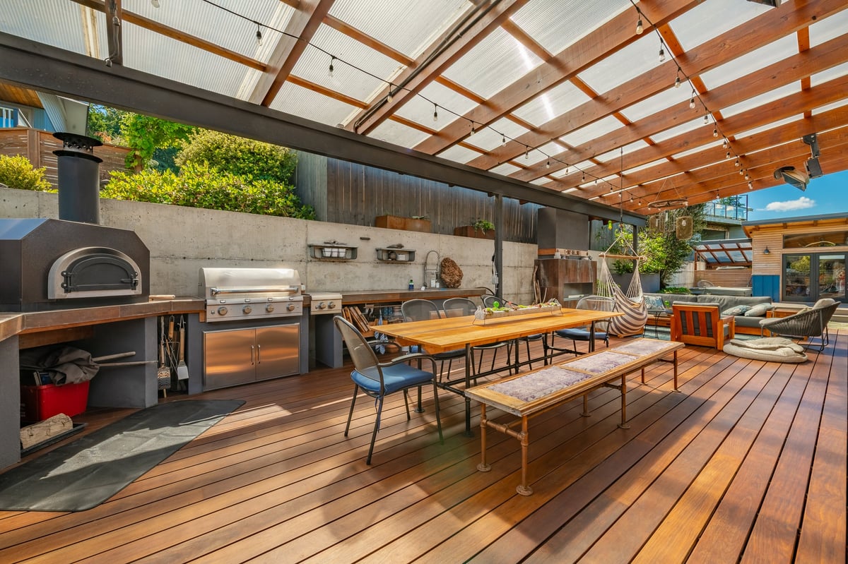 A large modern outdoor living space with a covered patio, built-in pizza oven, stainless steel grill and smoker, wooden decking, and dining and lounge areas