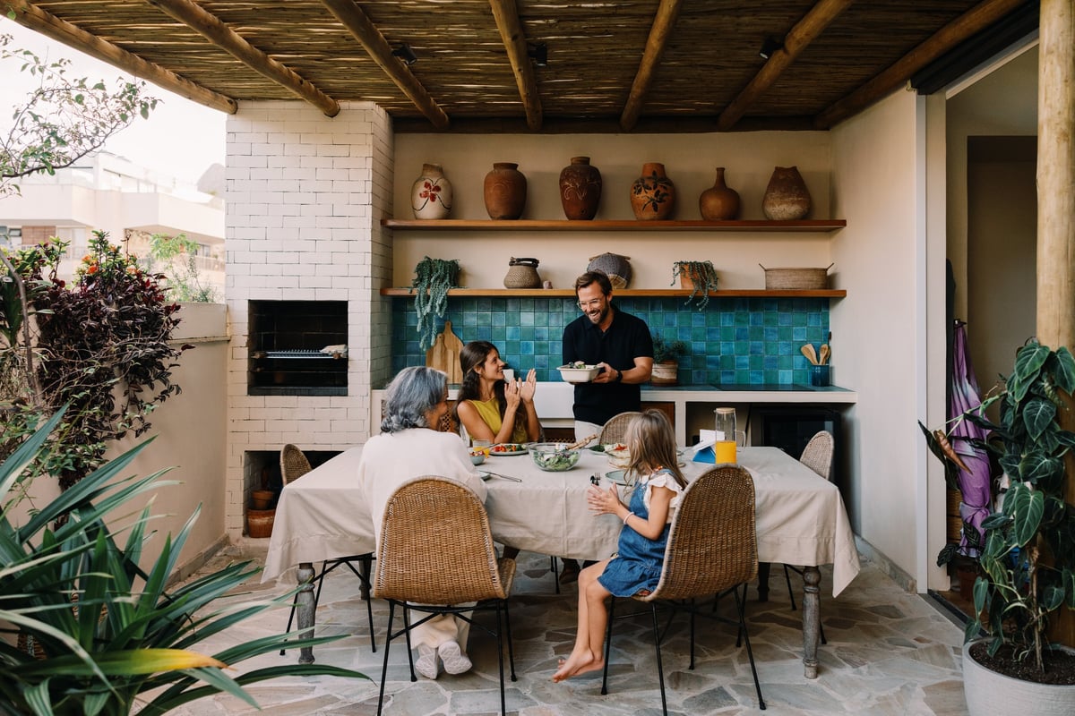 A family enjoys a casual outdoor meal on a covered patio. A man serves food while a woman and two children sit at a table, surrounded by plants and pottery.
