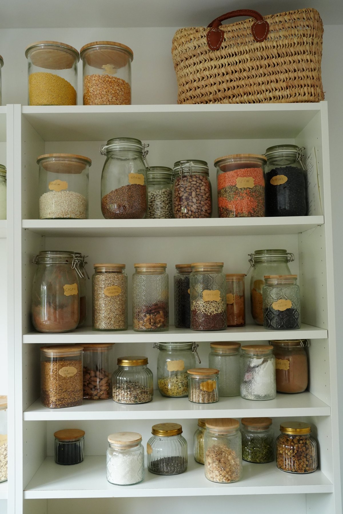 Pantry shelf filled with neatly arranged glass jars containing various grains, legumes, and spices. A woven basket sits atop, adding a rustic touch to this organized storage.