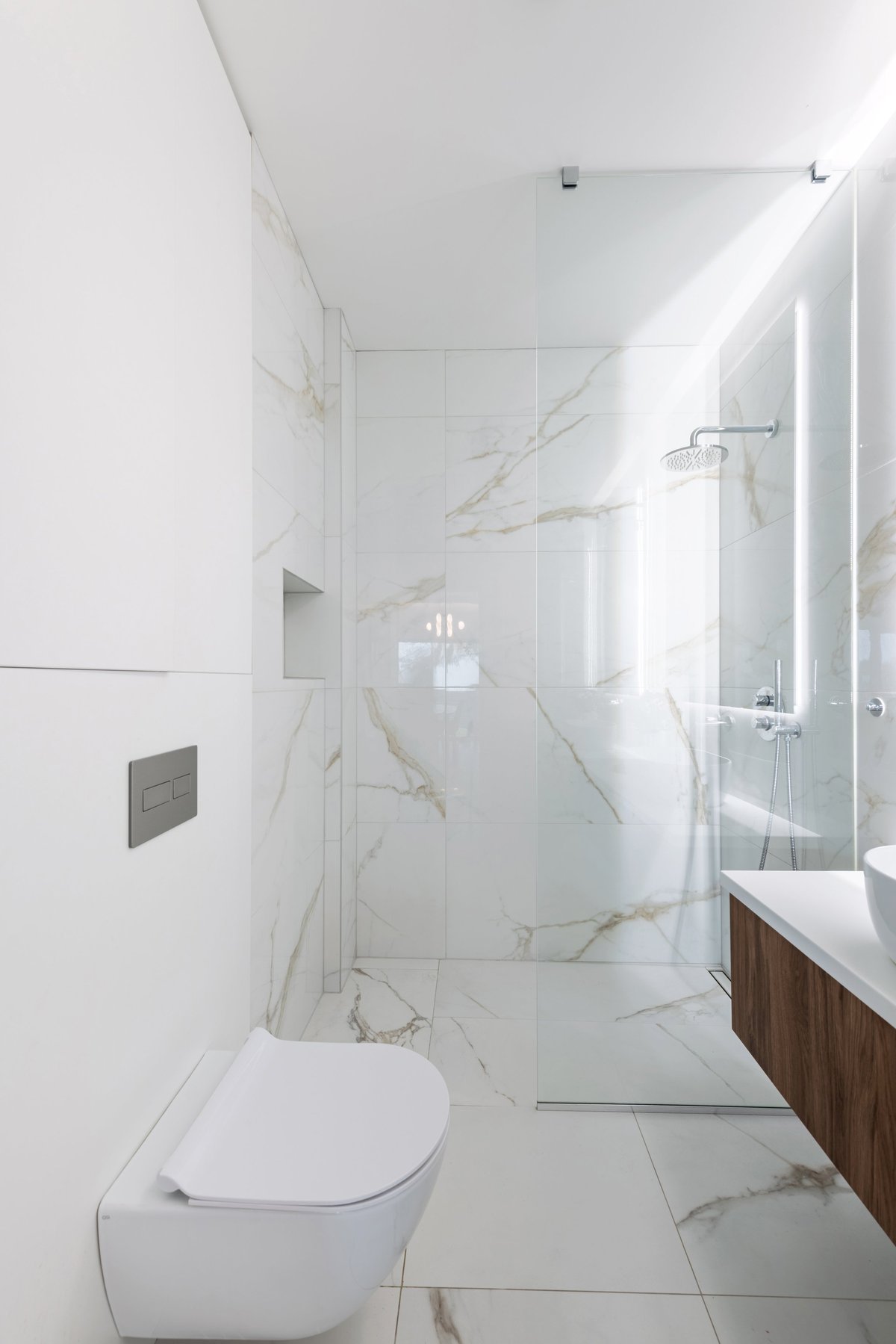 Bright modern bathroom with white marble tiles, frameless walk-in glass shower, wall-mounted toilet and floating wood vanity. Clean, minimalist design with soft natural light and luxurious, contemporary interior detailing