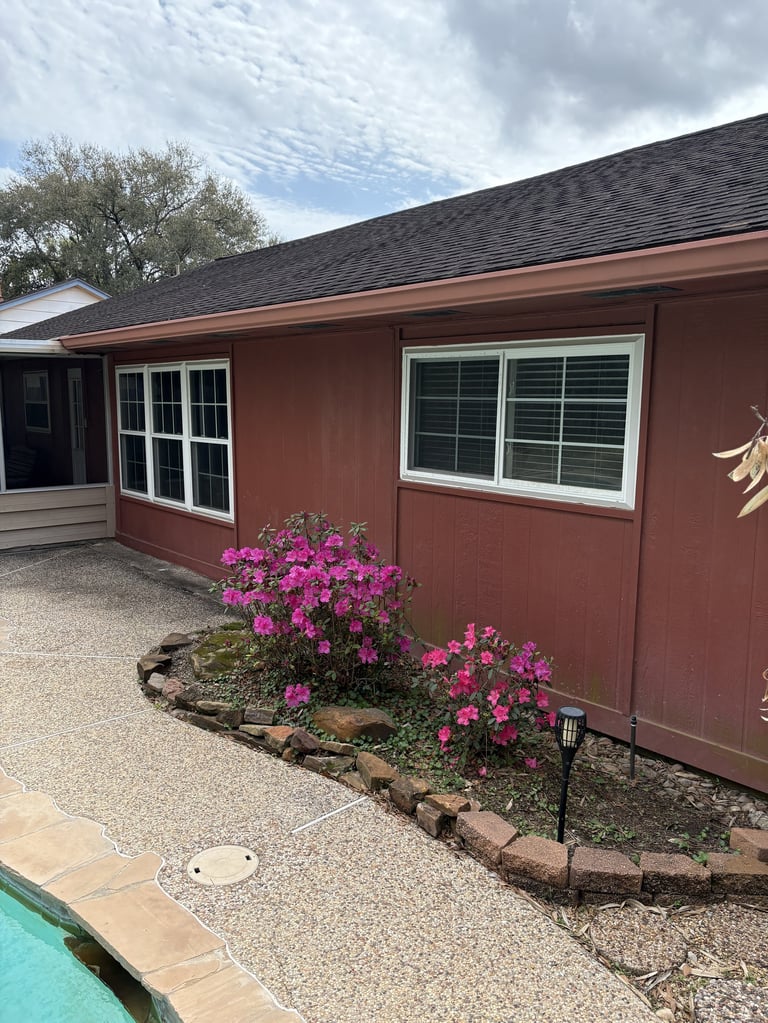 Red house with white-trimmed windows and gravel patio, featuring bright pink flowering shrubs in front landscaping