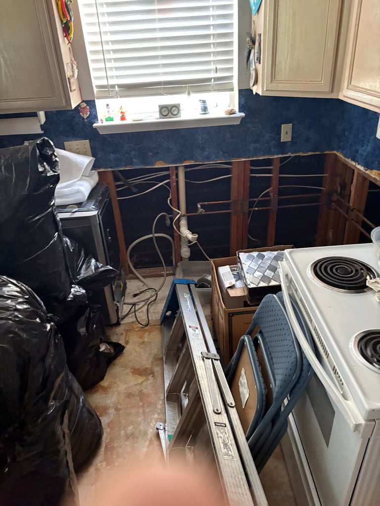Kitchen with blue walls during renovation, showing white stove, exposed wood frames, black plastic sheeting, and construction materials