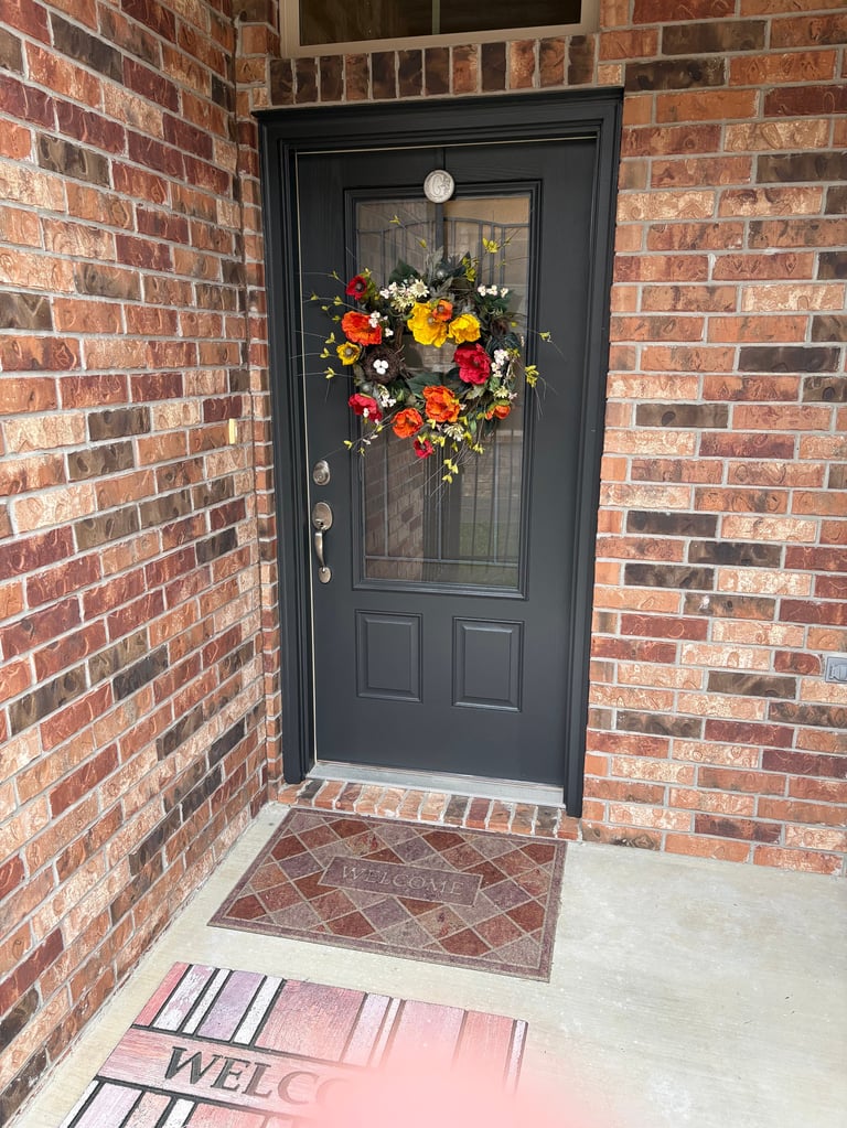 Black front door with colorful flower wreath on red brick home entrance with welcome mat - exterior rehab after