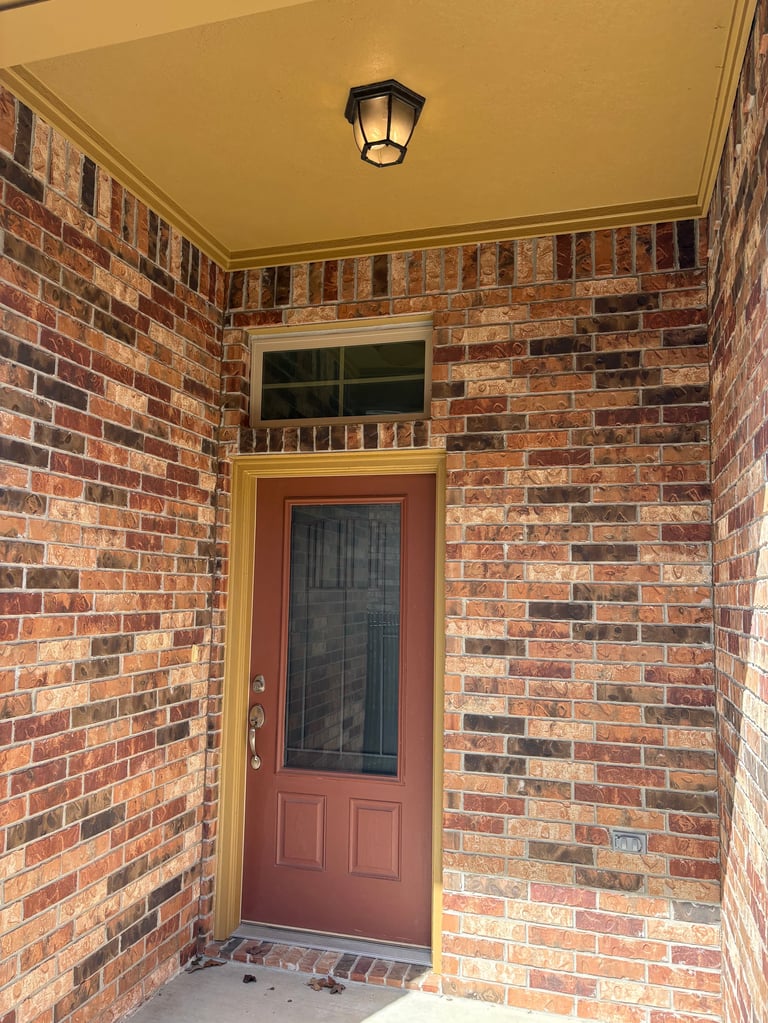 Red door with glass panel on brick entryway, yellow ceiling with black sconce light - exterior entryway before renovation