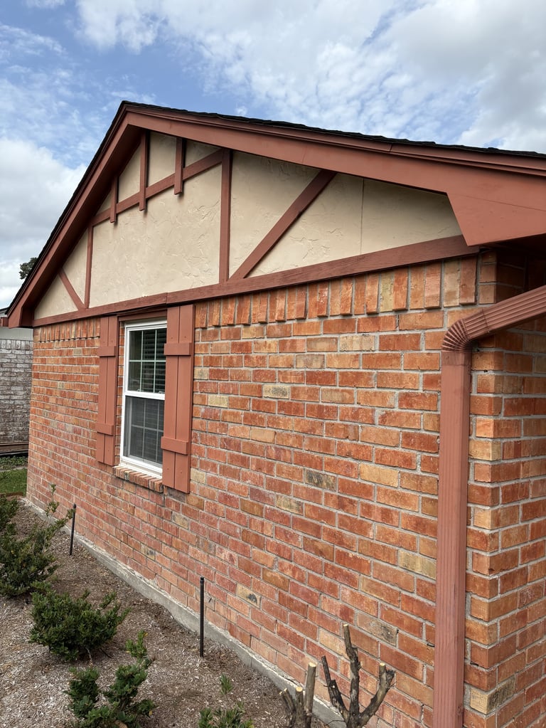 Brick cottage with red trim and decorative half-timbering on upper gable, white window, and manicured garden