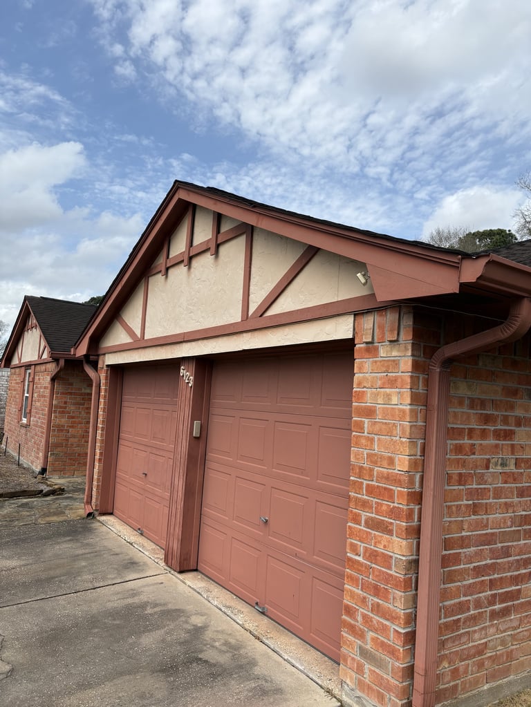 Two-car garage with red brick walls and red panel doors, featuring Tudor-style architectural details and trim under a partly cloudy sky