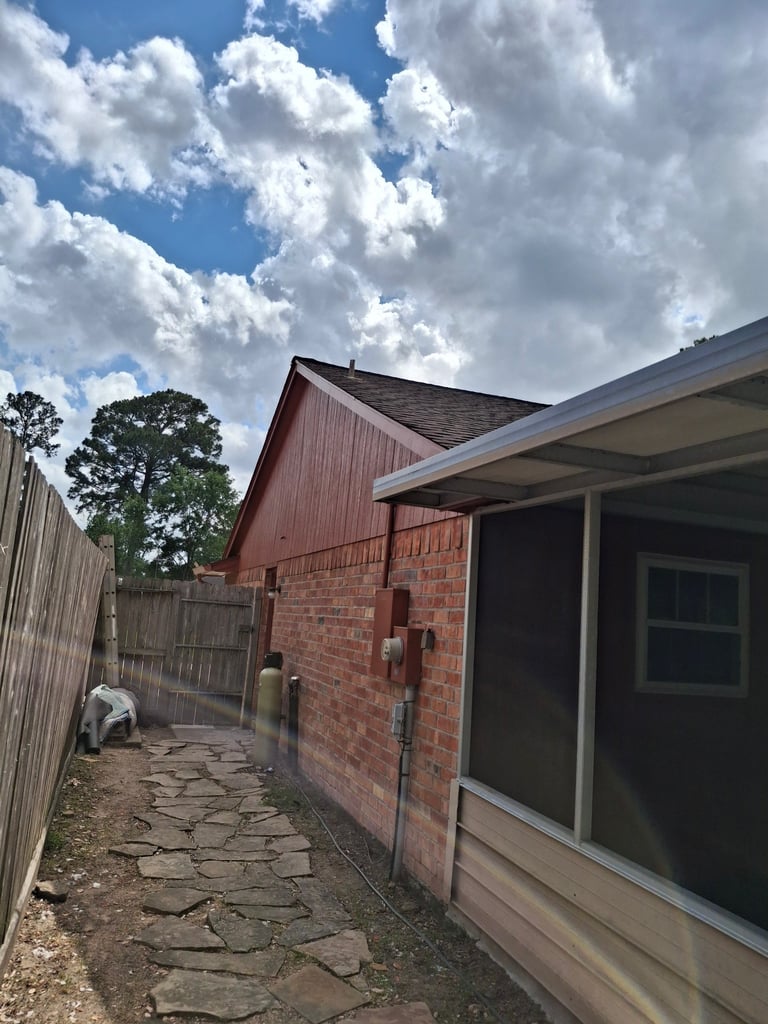 Brick building with red metal roof and modern extension, viewed from narrow stone pathway with metal fence and trees under blue sky with white clouds